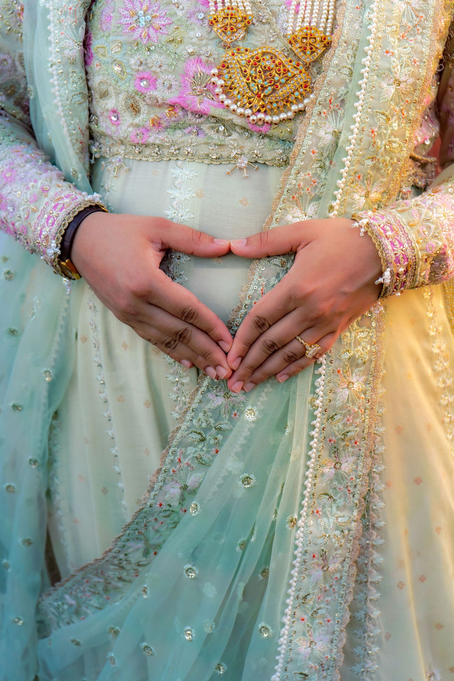 Couple making heart shape with hands over mint green embroidered Indian bridal lehenga with gold jewelry.
