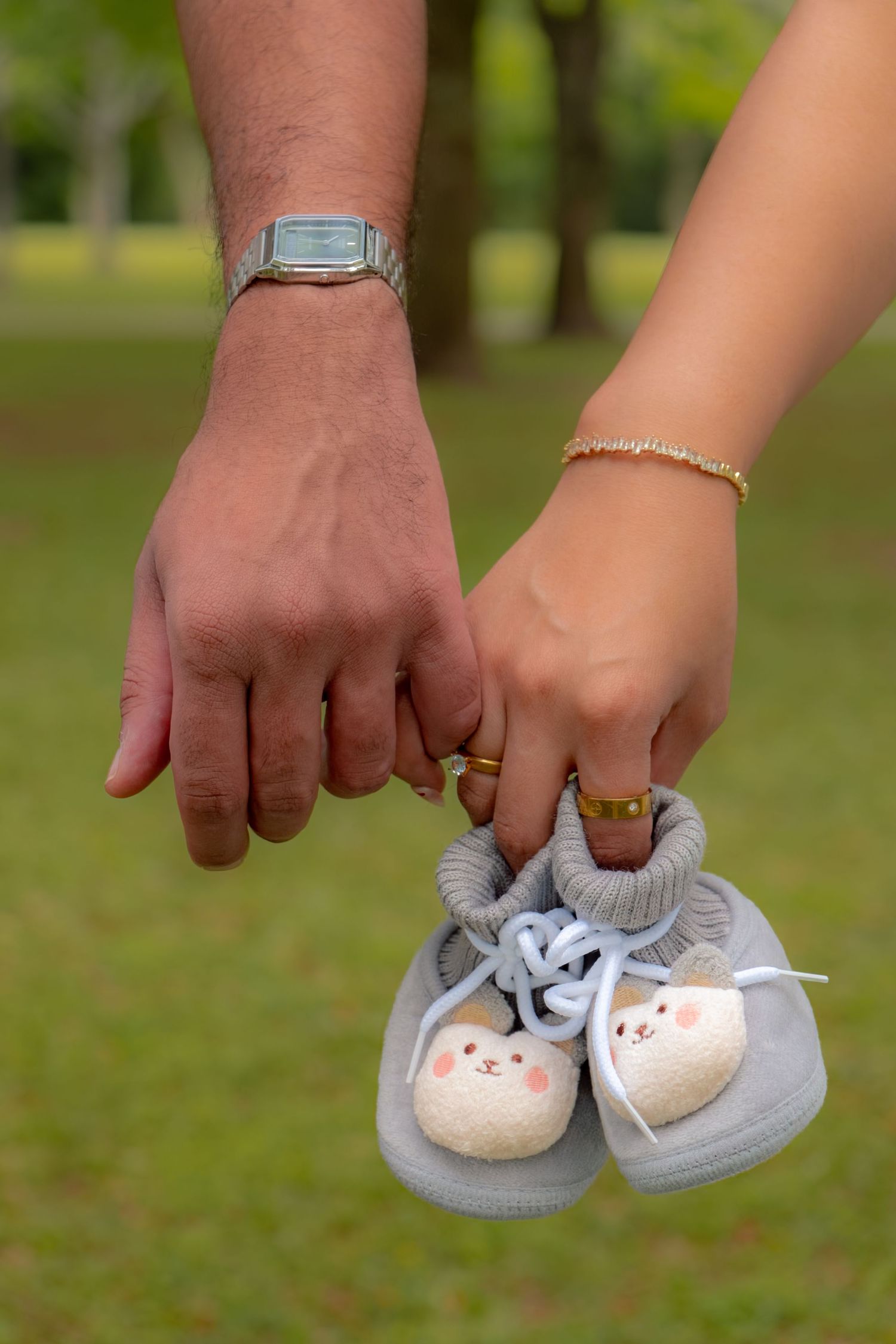 Couple holding tiny gray baby booties together outdoors, wearing wedding rings and accessories.