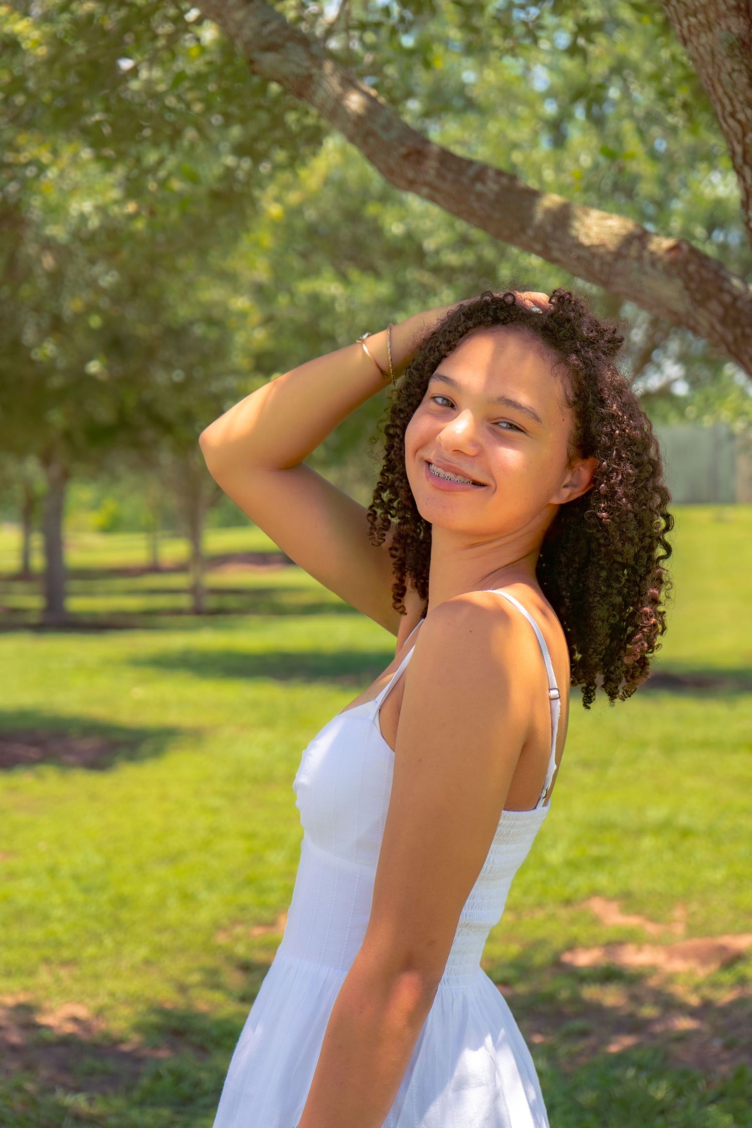 Young woman with curly hair in white sundress smiling outdoors in sunny green park setting.