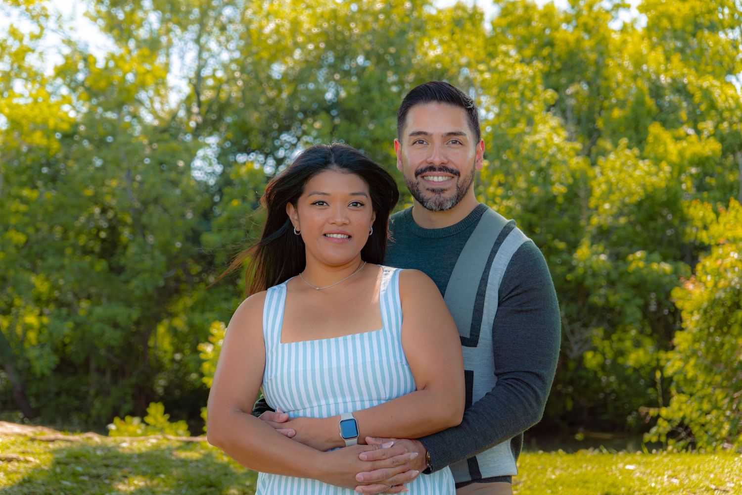 A smiling couple poses outdoors in a sunny park surrounded by lush green trees on a bright day.