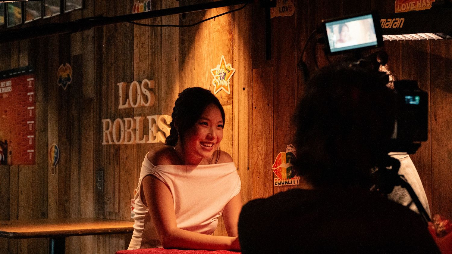 Smiling woman in white dress being interviewed on camera at Los Robles rustic wooden backdrop.