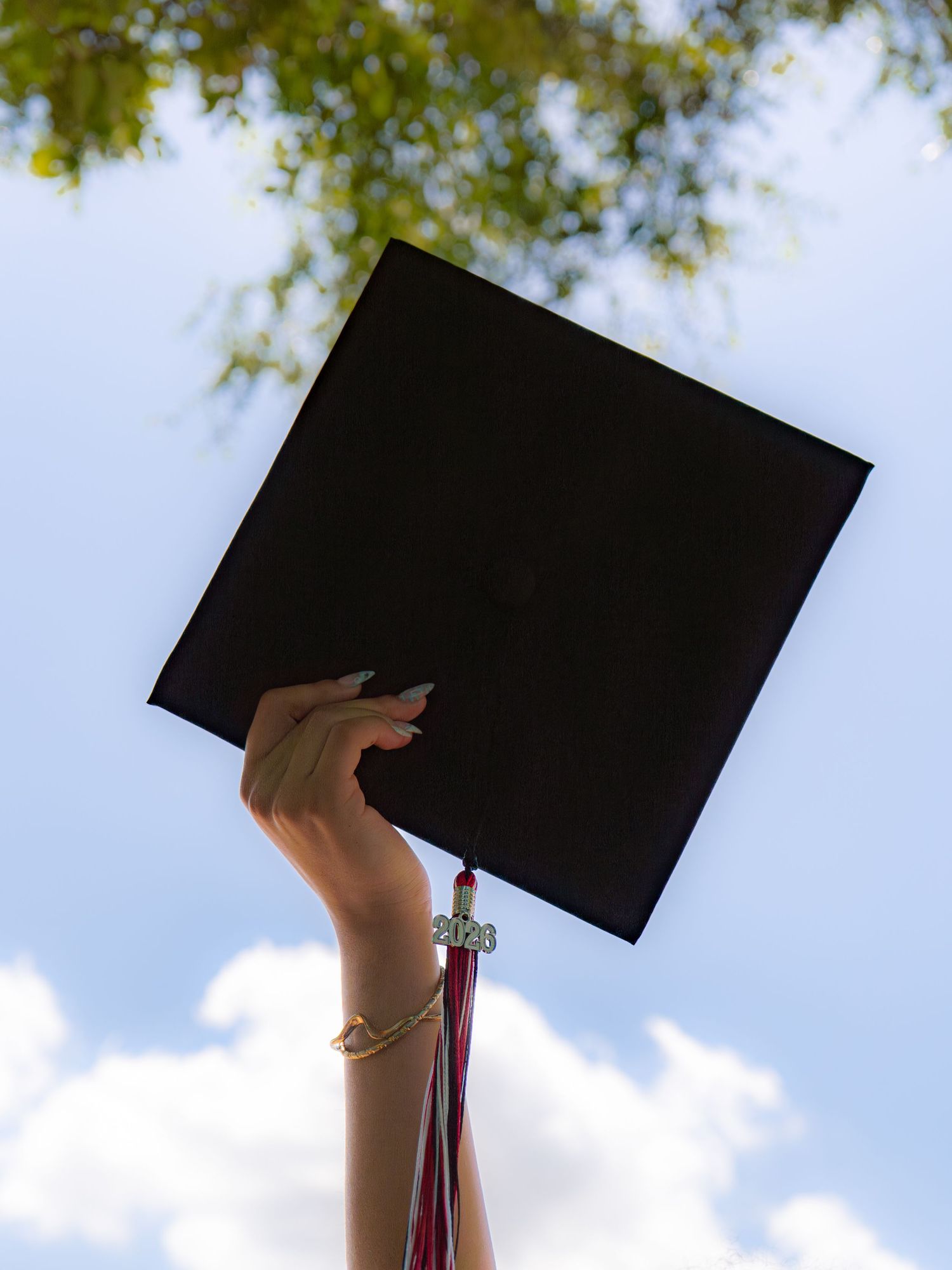 A hand raised holding a black graduation cap against a blue sky with green tree branches overhead.
