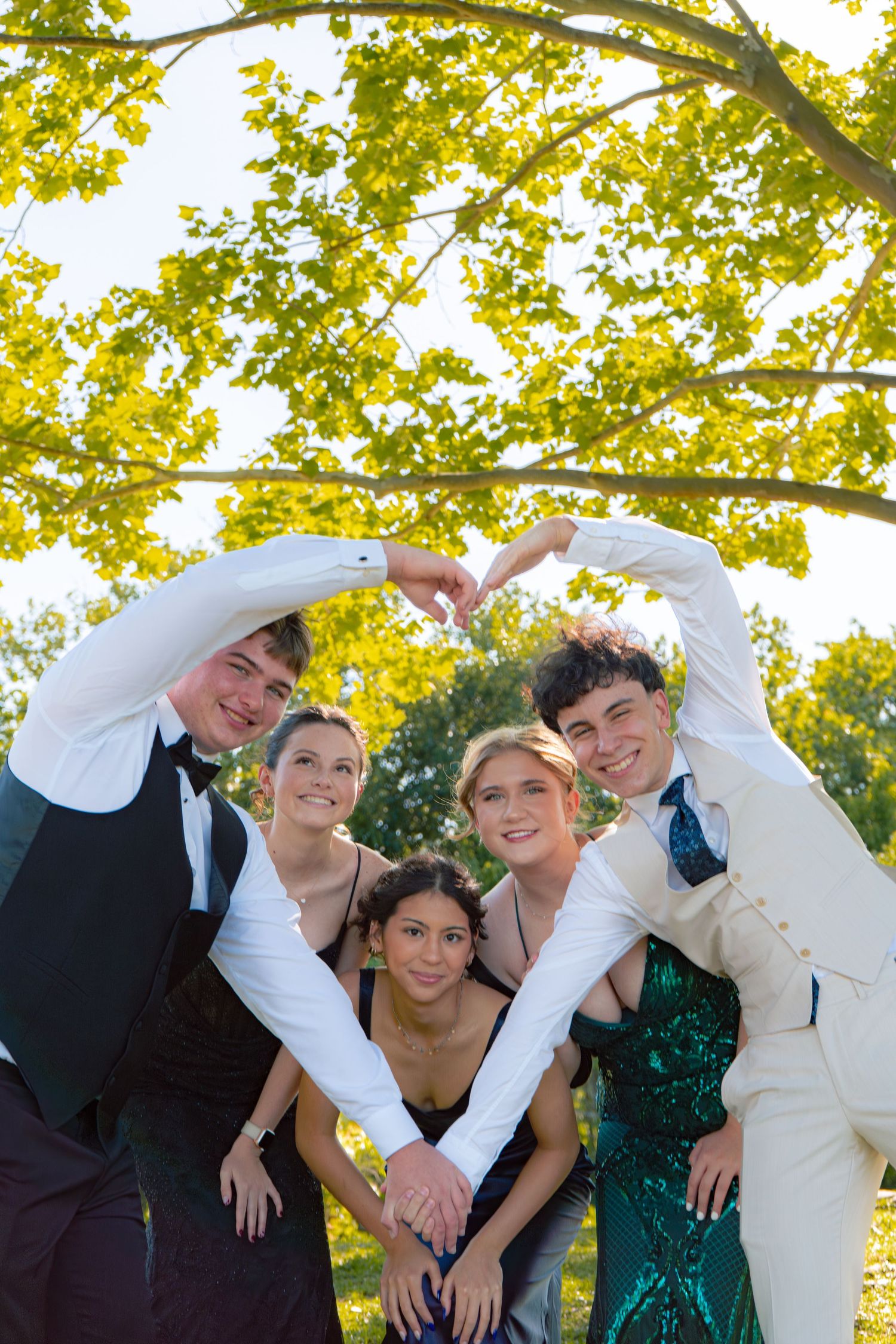 Five prom-dressed teens form heart shape with hands under leafy tree.