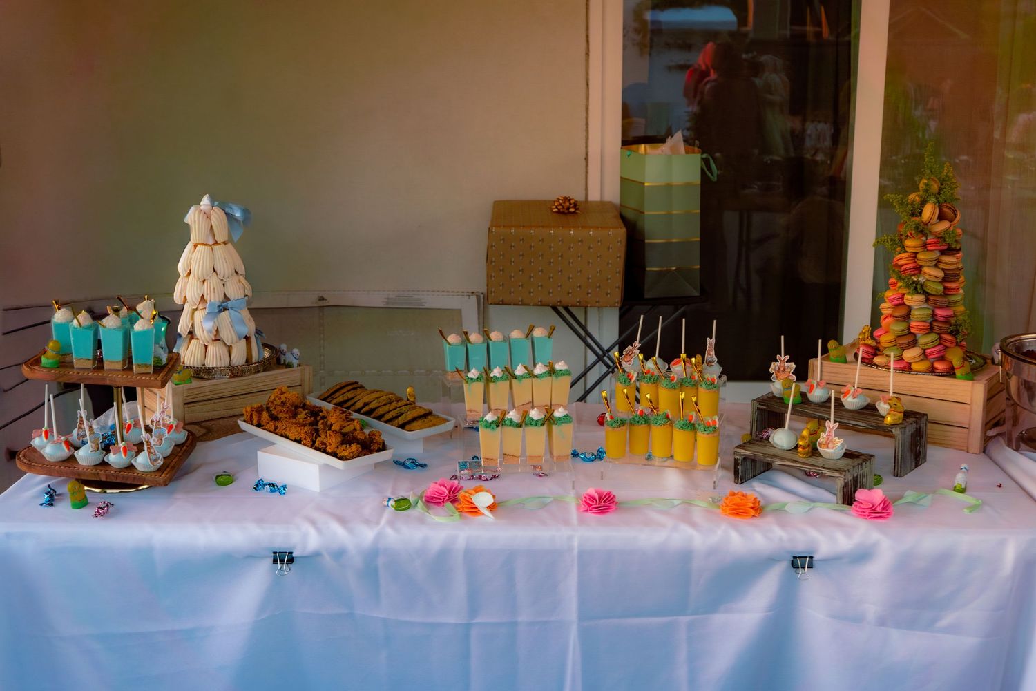 Elegant dessert table with macarons, cake pops, yellow drinks, and colorful treats on a white tablecloth.
