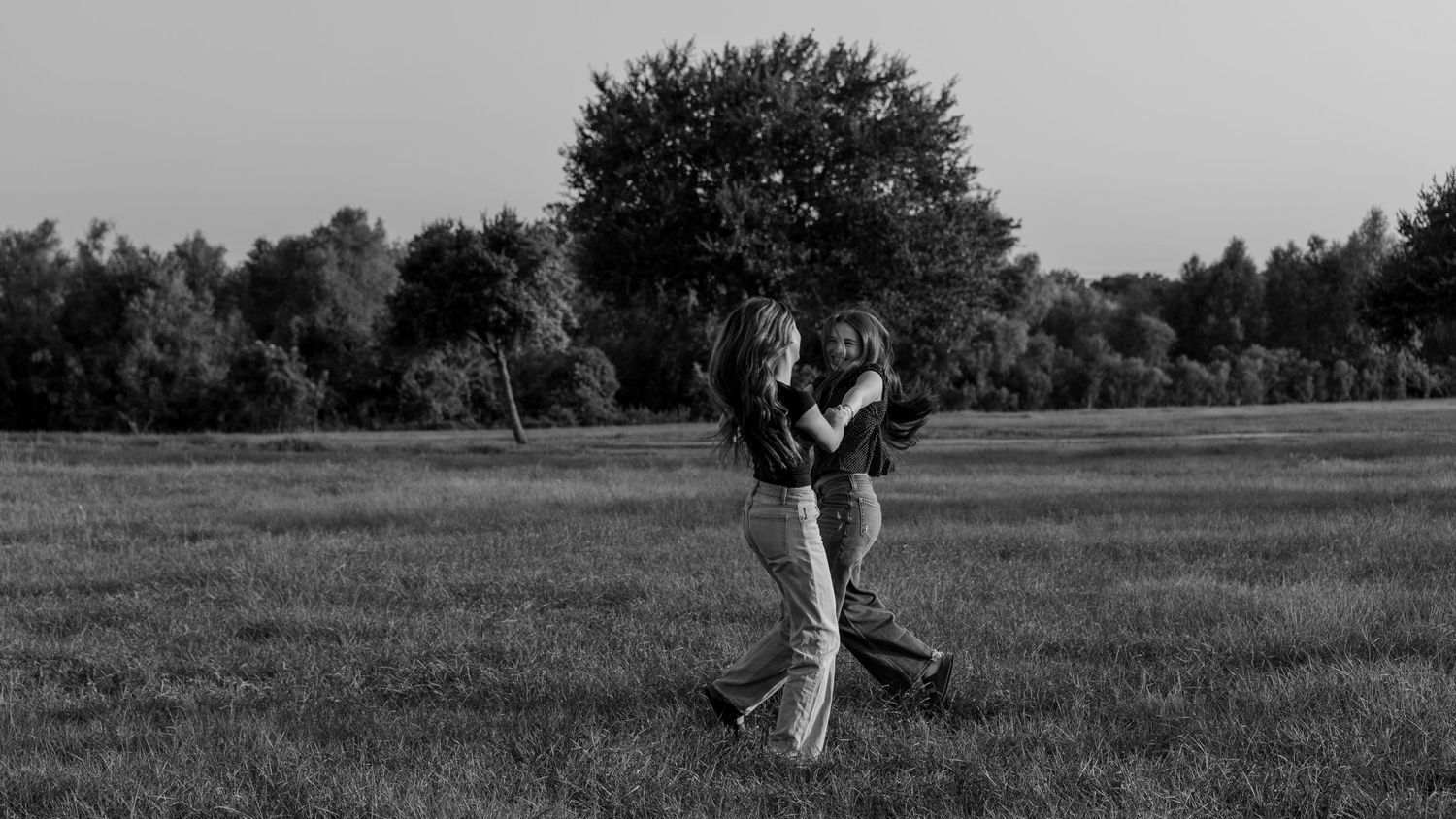 Two women embrace and dance together in a scenic open field with trees in the background in a black and white photo.