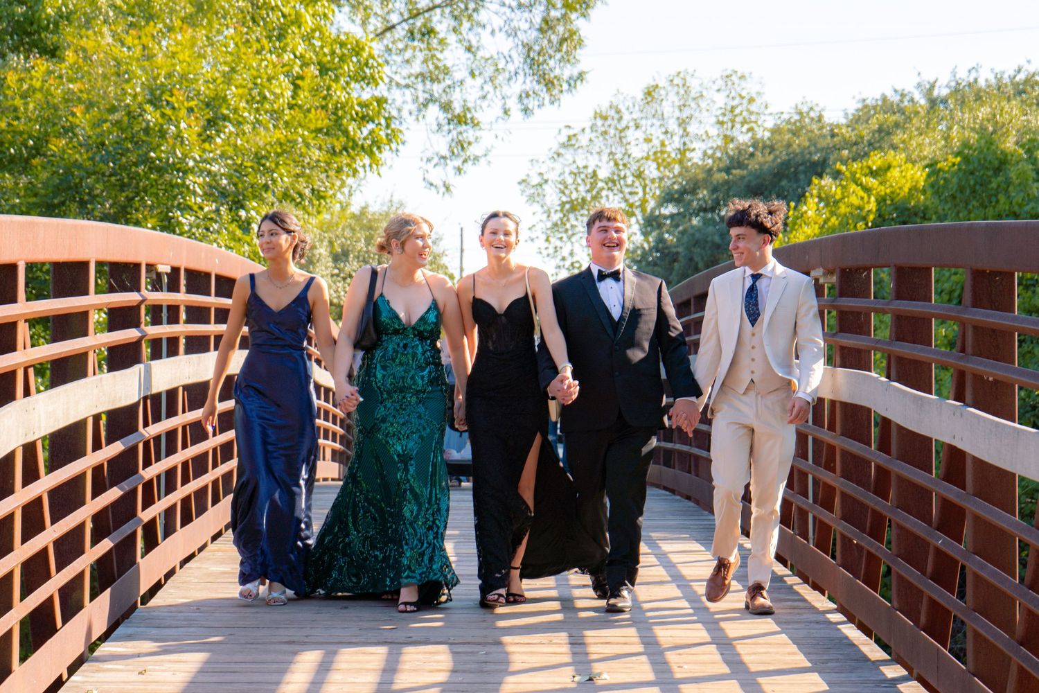 Five teenagers dressed in elegant prom attire walk together across a wooden bridge surrounded by green trees.