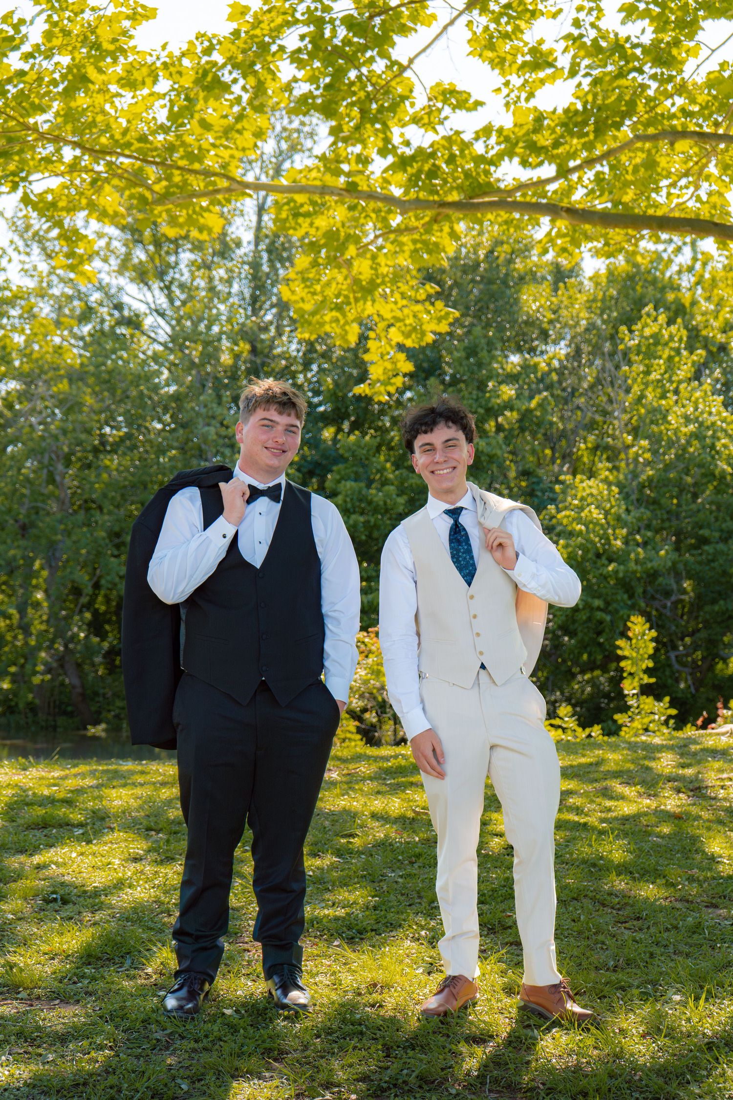 Two young men in formal suits pose outdoors under yellow-green tree branches on a sunny day.