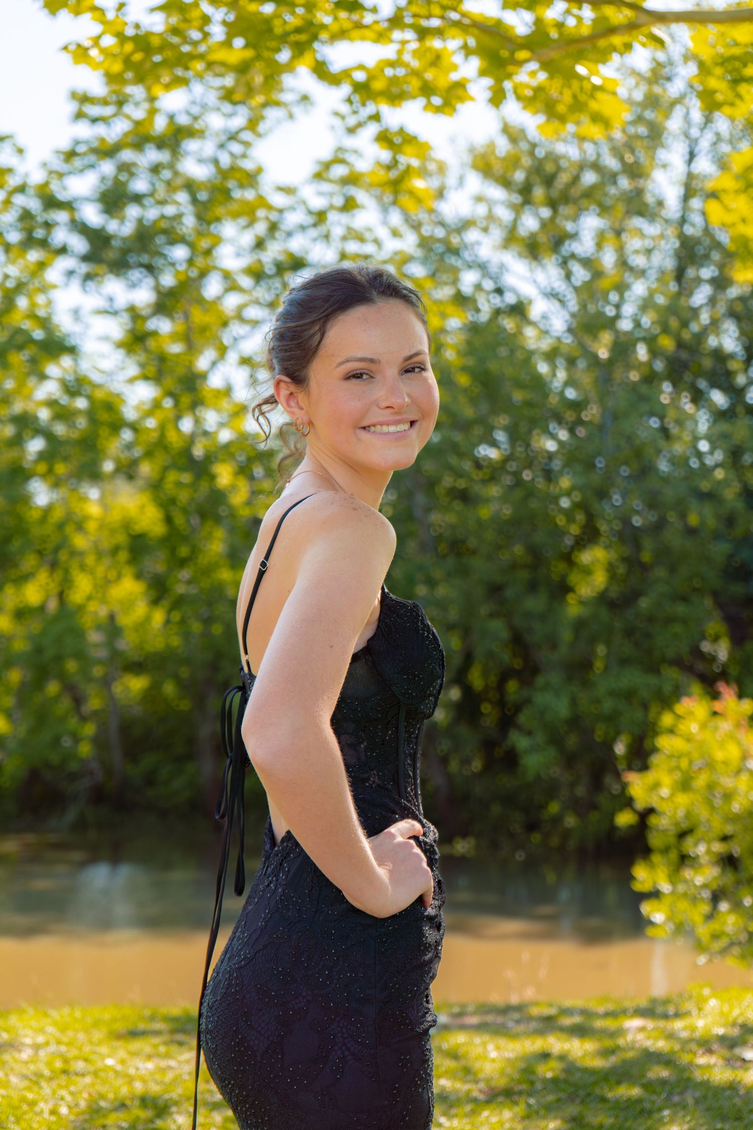 Smiling teenage girl in backless black sequin prom dress poses outdoors near a sunlit pond.