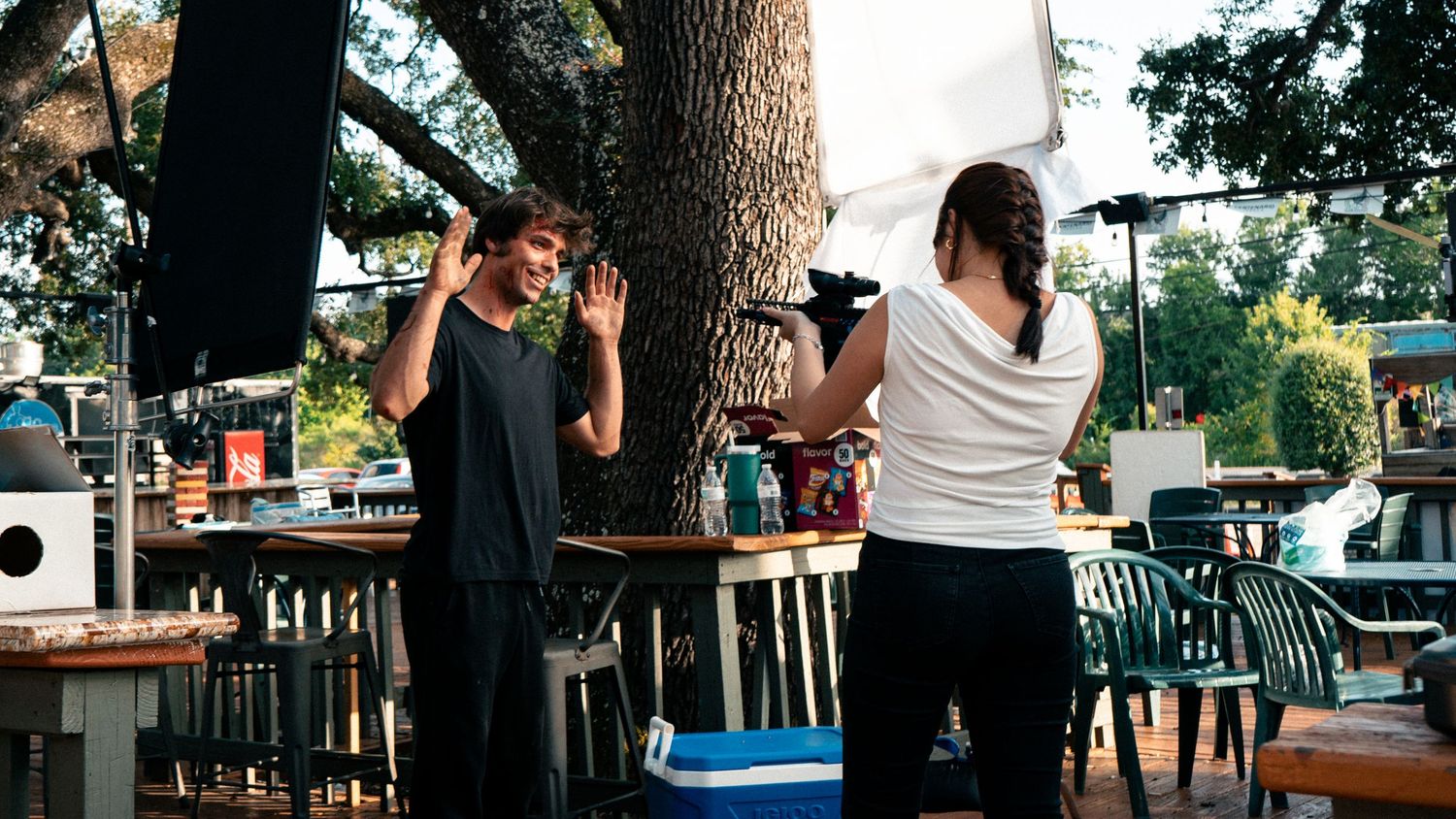 Two people interact near a tree in an outdoor restaurant patio setting on a sunny day.