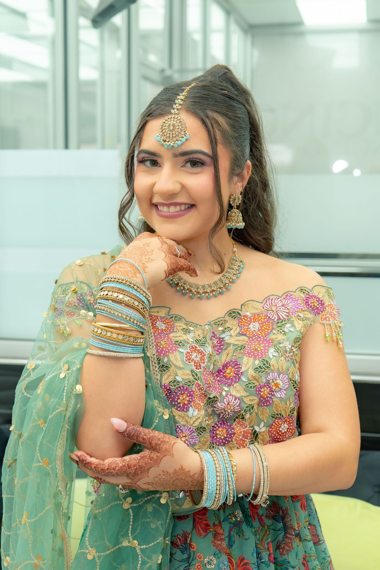 Young woman in teal floral lehenga with gold jewelry, bangles, and mehndi, smiling at camera.