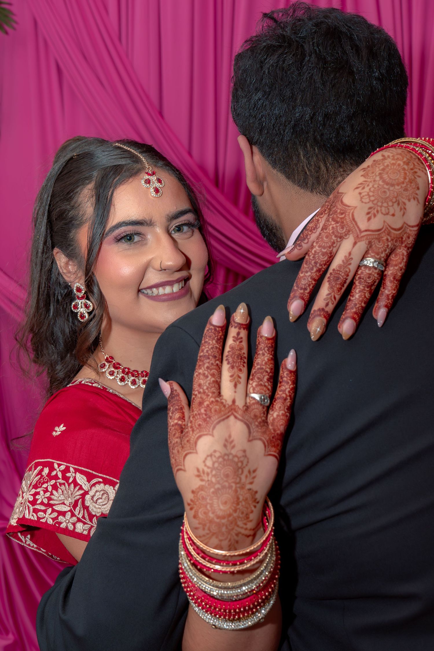 Indian bride in red lehenga with intricate mehndi embracing groom in black suit at wedding.