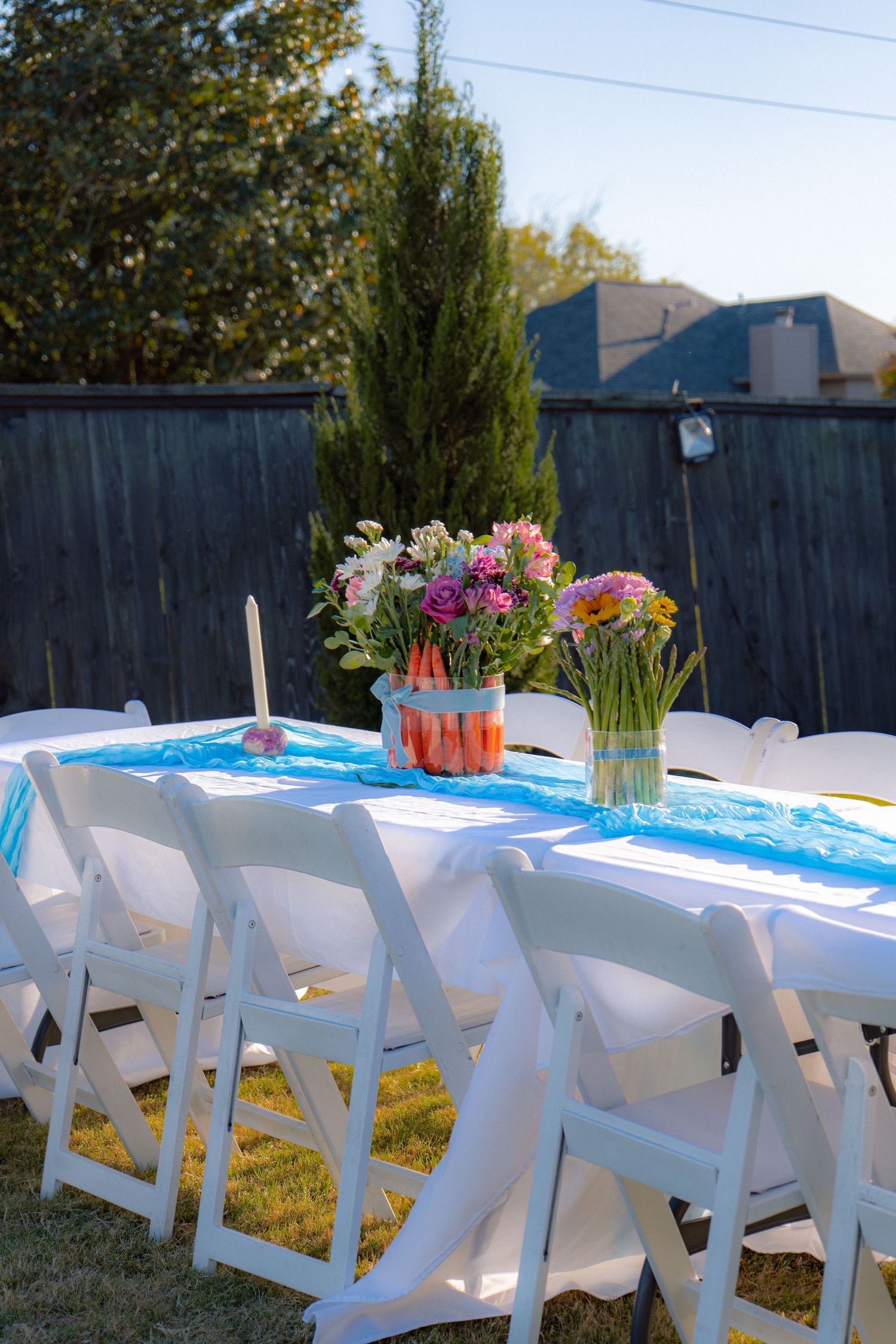 Outdoor baby shower reception table with white folding chairs, blue runner, and colorful floral centerpieces in a backyard.