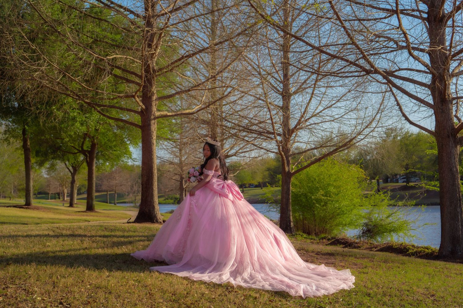 Young woman in a pink ball gown quinceañera dress poses gracefully in a park with bare trees near a lake.