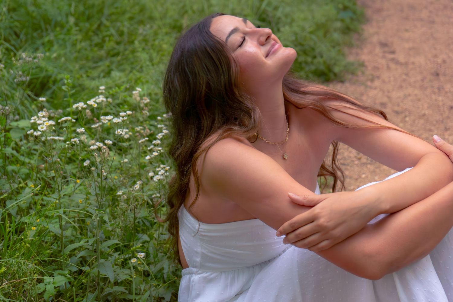 Young woman in white dress sitting outdoors near wildflowers with eyes closed.
