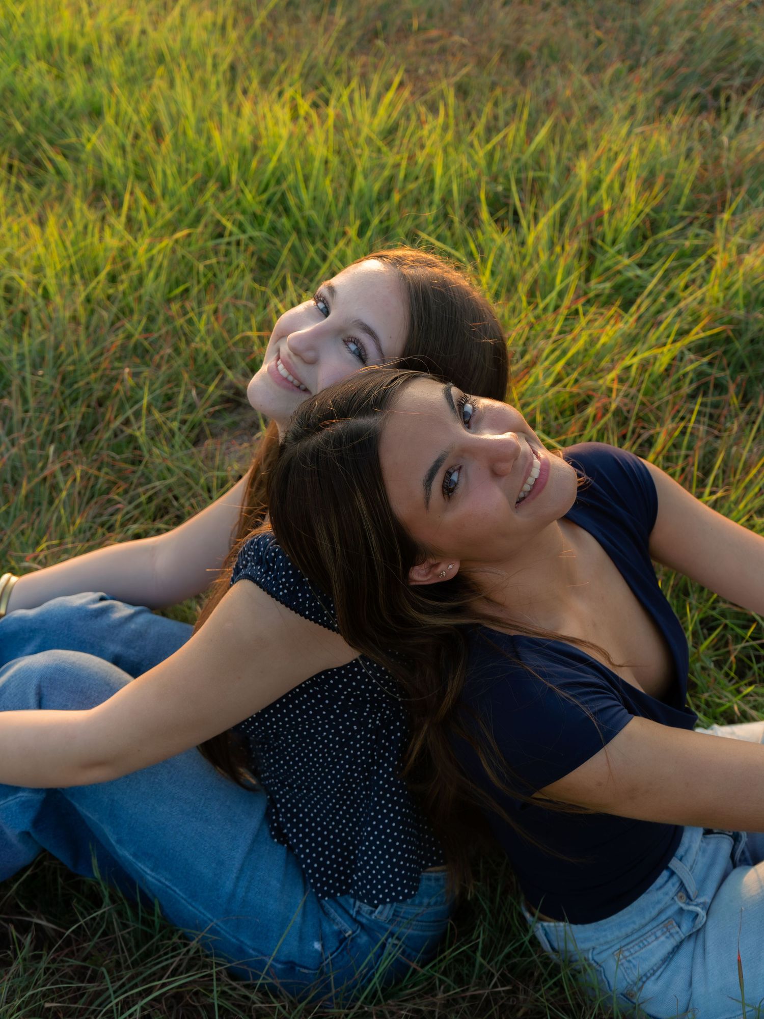 Two young women sitting back-to-back on green grass field, smiling up at camera in warm golden hour light.