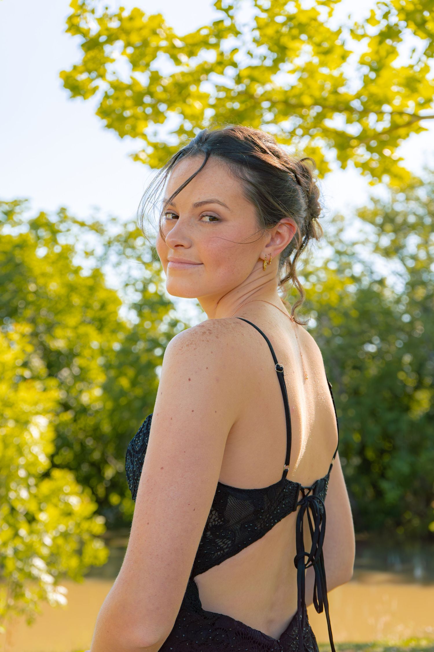 Young woman in elegant backless black lace dress poses outdoors with lush green trees in background.