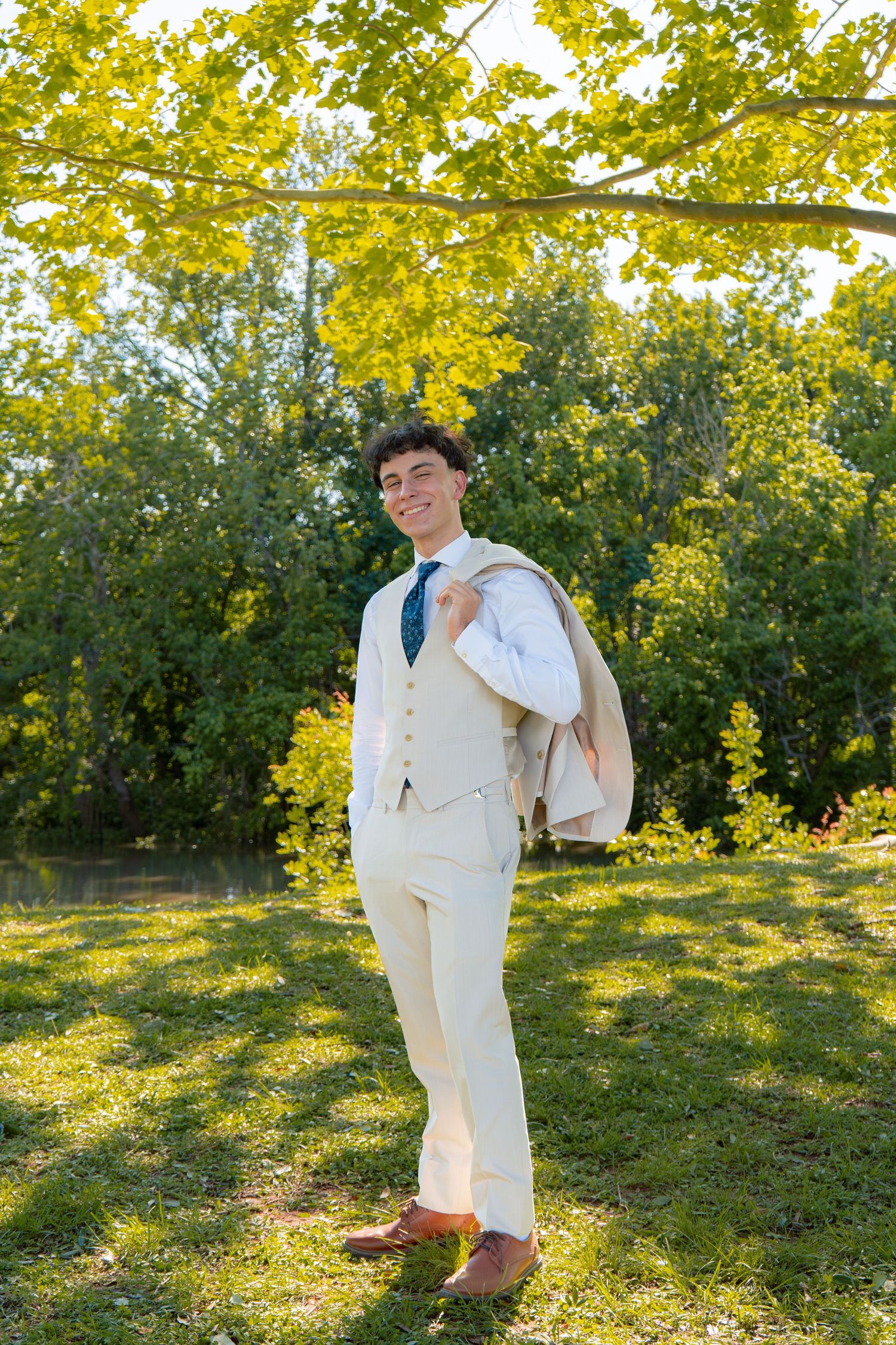 Smiling young man in a cream vest and trousers carries his suit jacket over his shoulder in a sunny park setting.