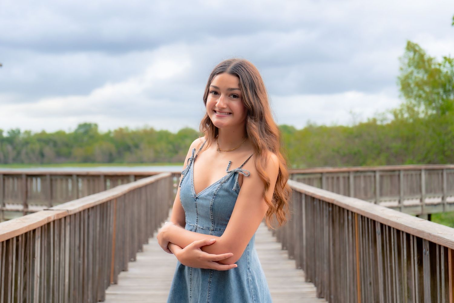 Young woman in blue dress smiling on wooden bridge boardwalk surrounded by lush greenery.