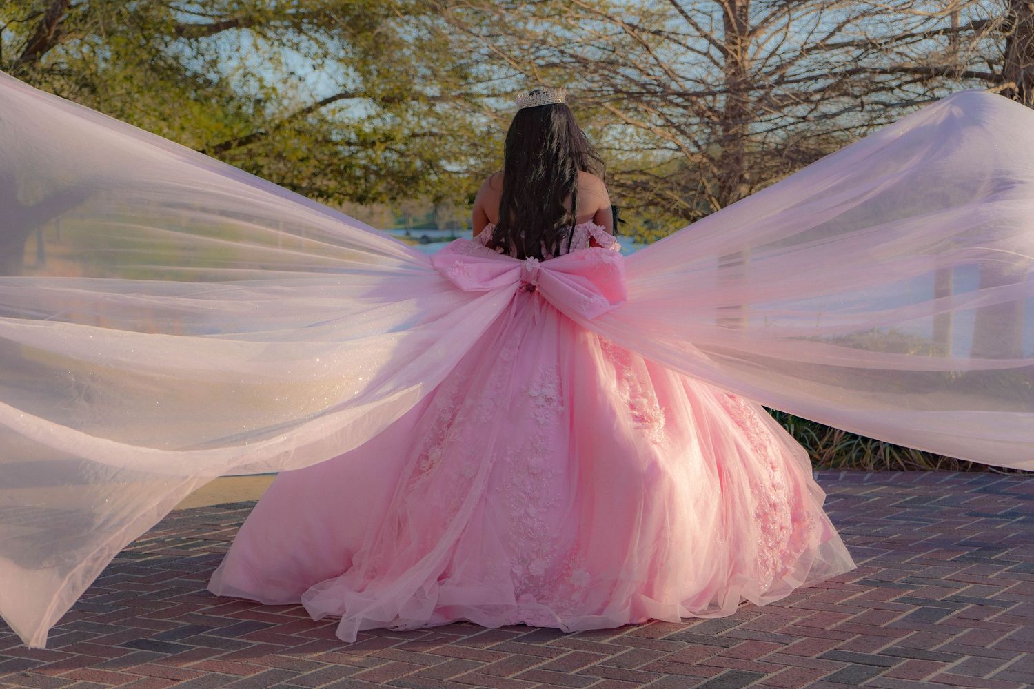 Young woman in a pink ball gown with flowing cape twirls outdoors, showcasing an elegant quinceañera dress.