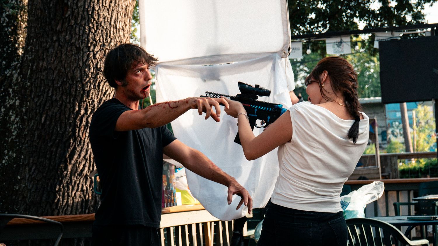 Two people engage in a staged combat scene outdoors, with a man and woman handling a prop rifle near a tree.