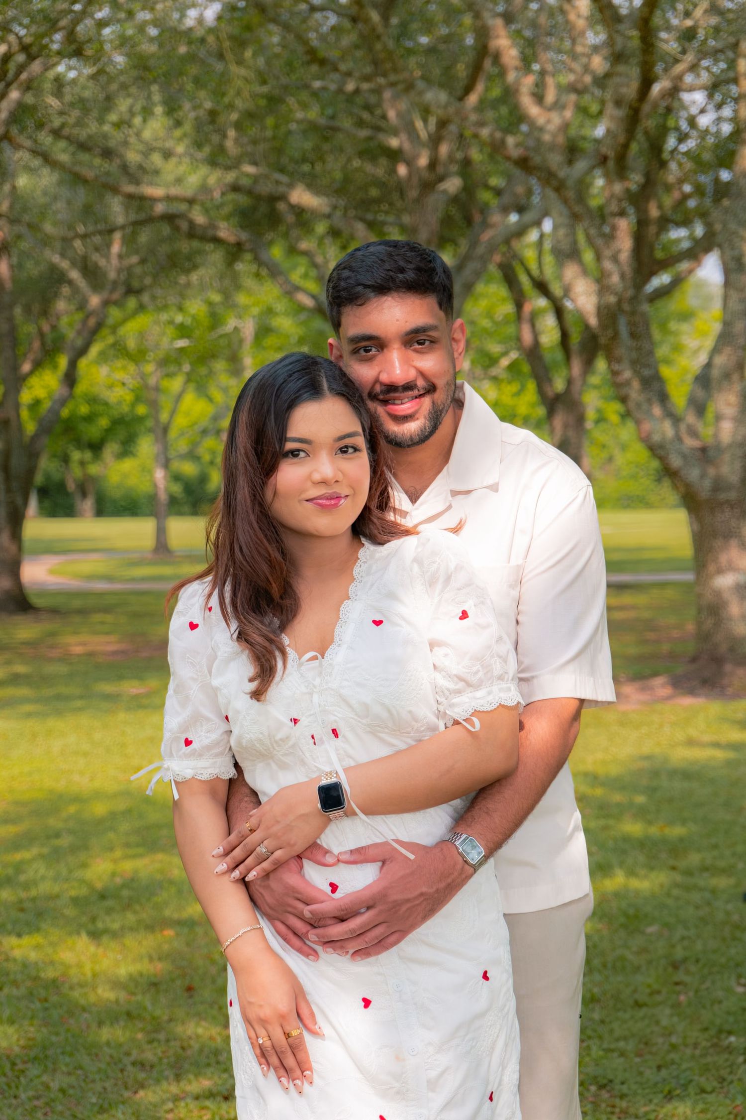 Couple in white outfits poses in a park, man embracing pregnant woman from behind, surrounded by lush green trees.