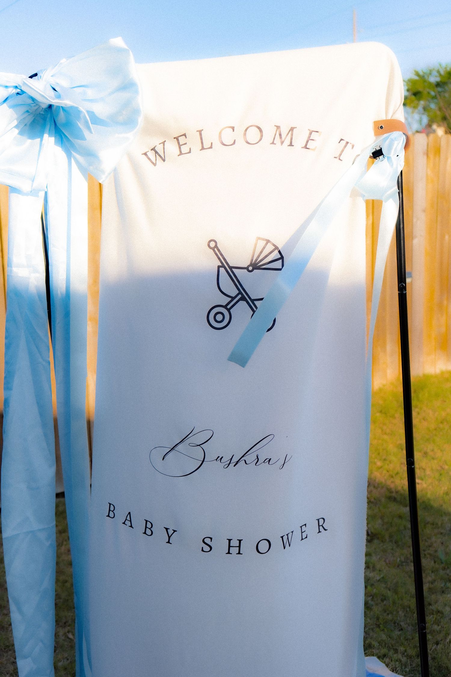 Welcome to Buskra's Baby Shower banner with blue ribbons and baby stroller illustration at an outdoor celebration.