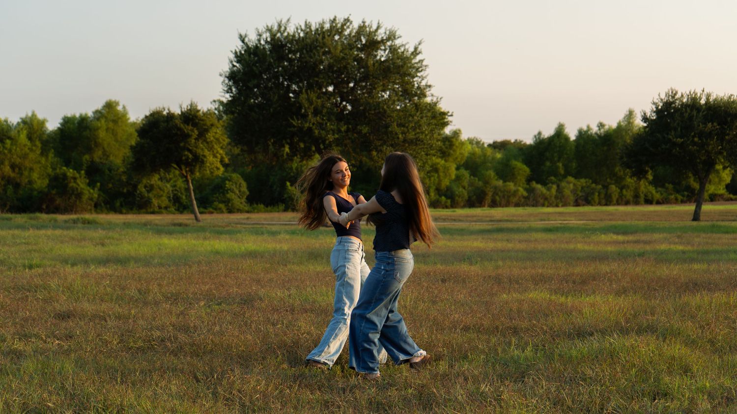 Two young women dancing together in a green meadow at sunset with trees in the background.