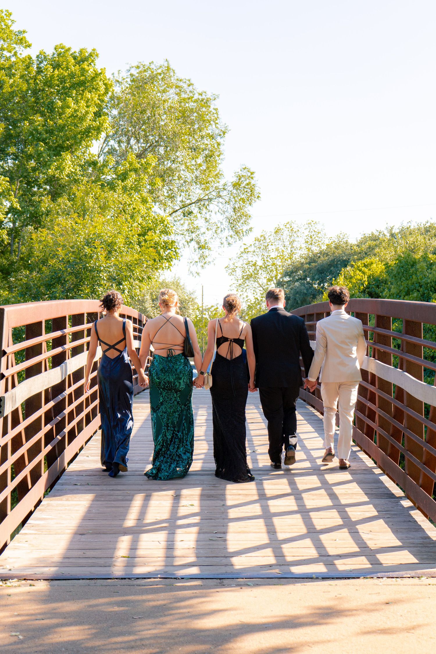 Group of prom-dressed teens walking away on wooden bridge surrounded by lush green trees.