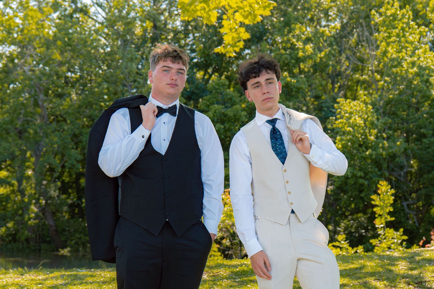 Two teenage boys in formal suits pose confidently outdoors with yellow-green foliage in the background.