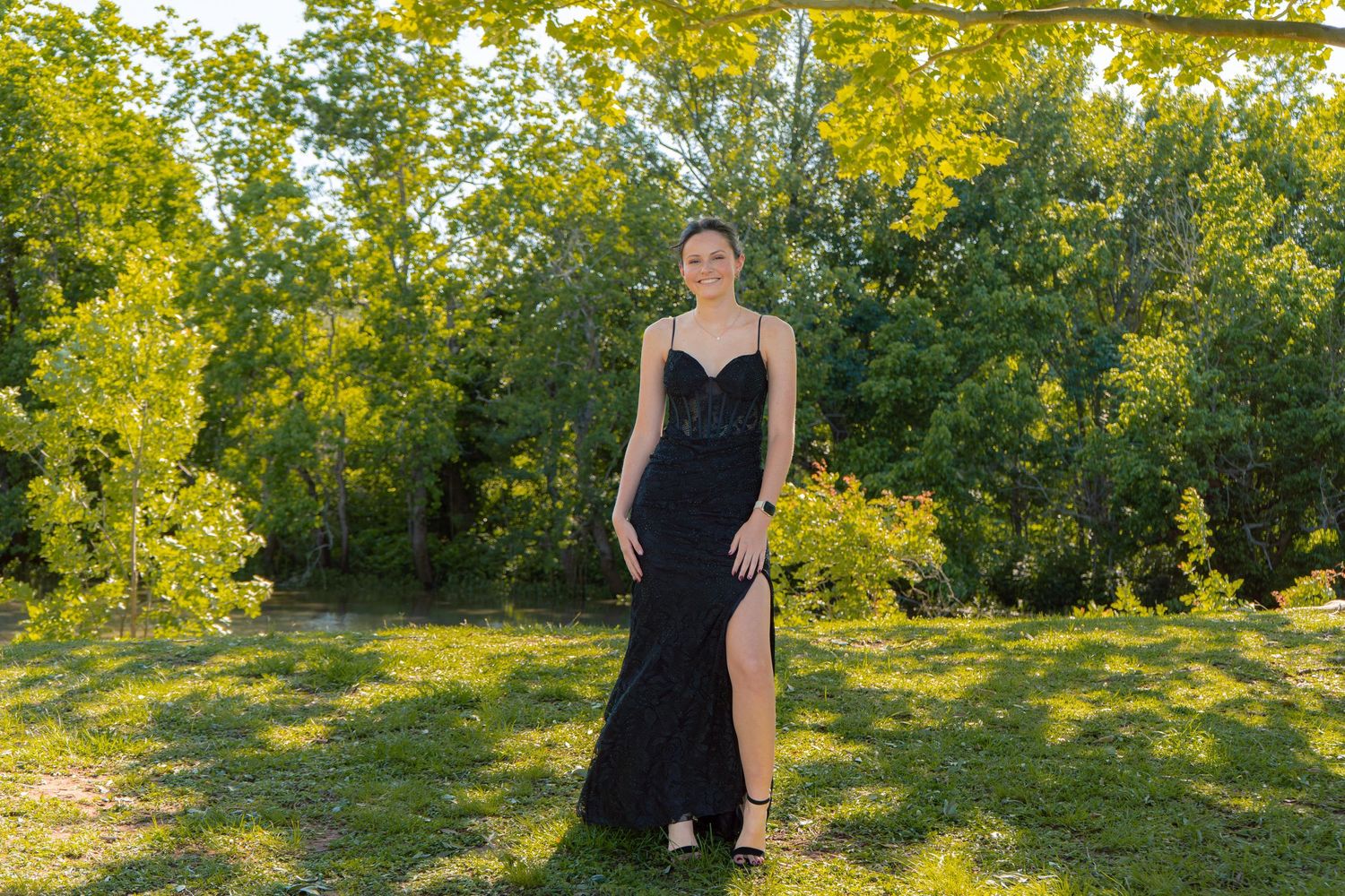 Young woman in a black spaghetti-strap formal gown with a slit poses gracefully in a lush green outdoor setting.