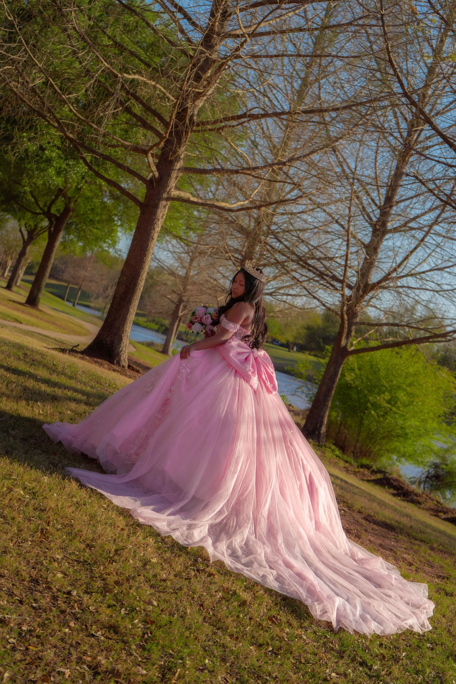 Young woman in elegant pink ball gown with floral bouquet poses gracefully among trees near a serene lake.