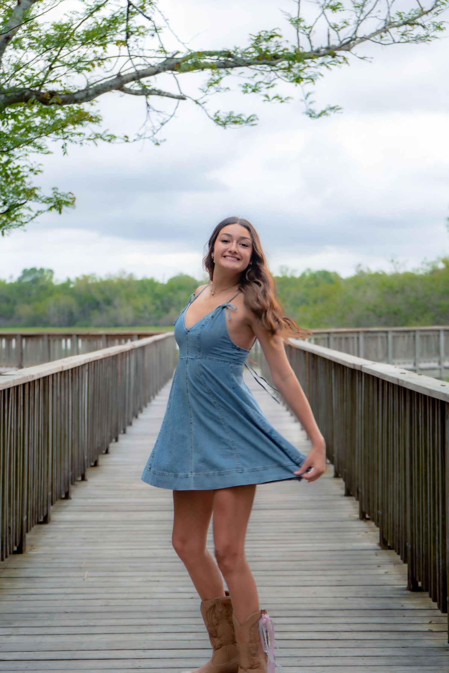 Young woman in blue denim dress and cowboy boots posing on wooden bridge.