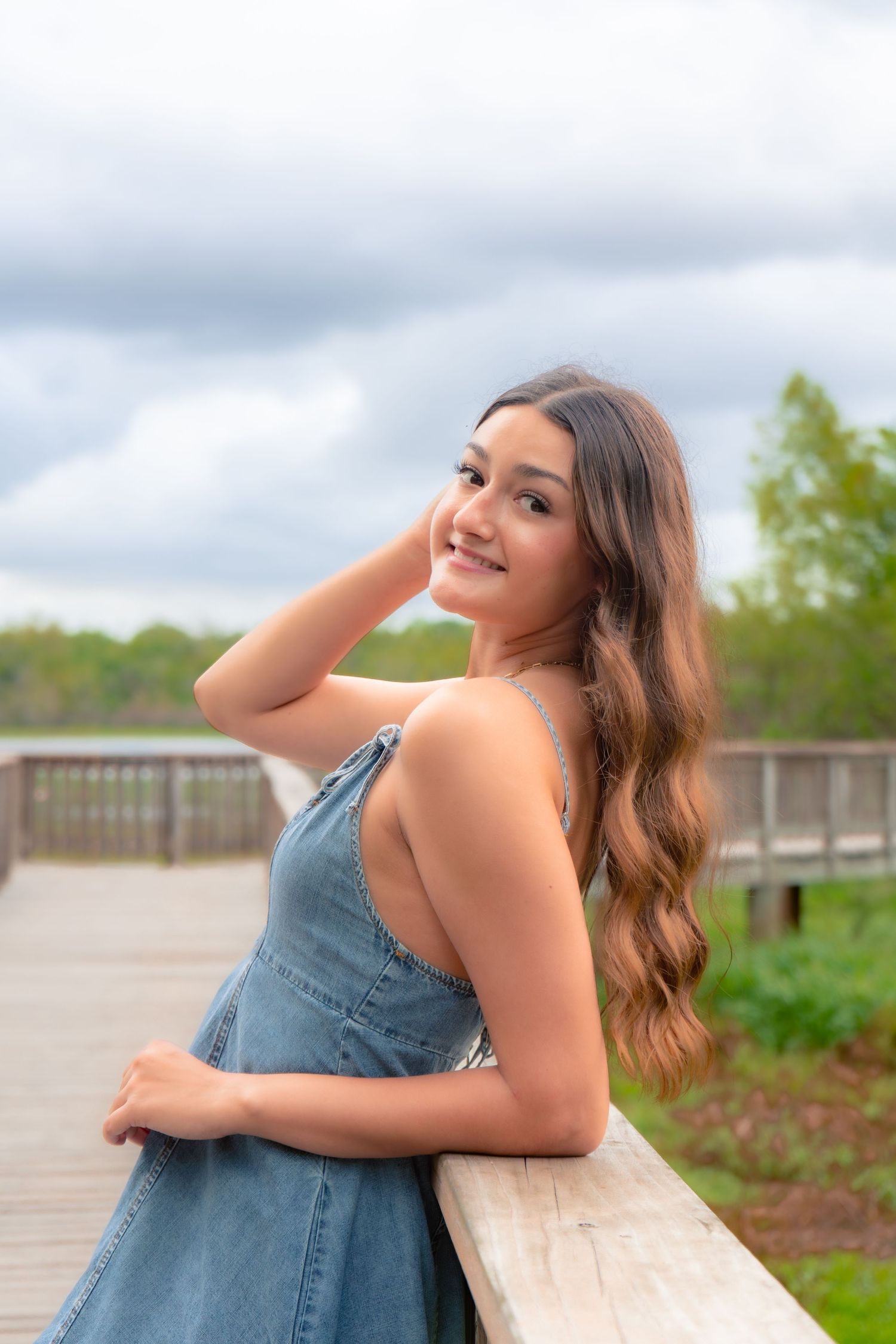 Smiling young woman in denim dress leaning on wooden railing of boardwalk near lake.