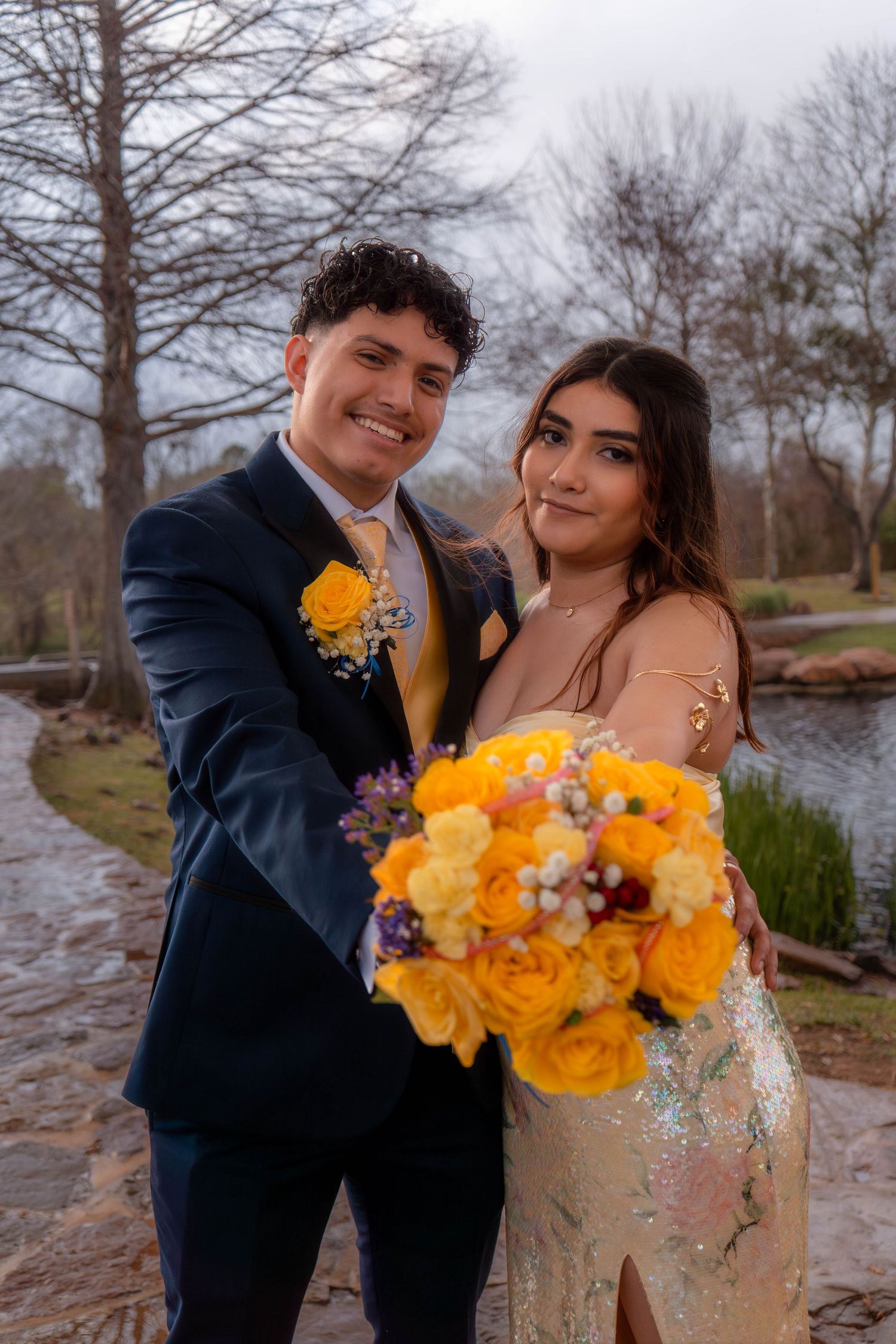 Smiling prom couple embracing outdoors, young man in navy tuxedo presenting yellow rose bouquet with girl in gold sequin gown.