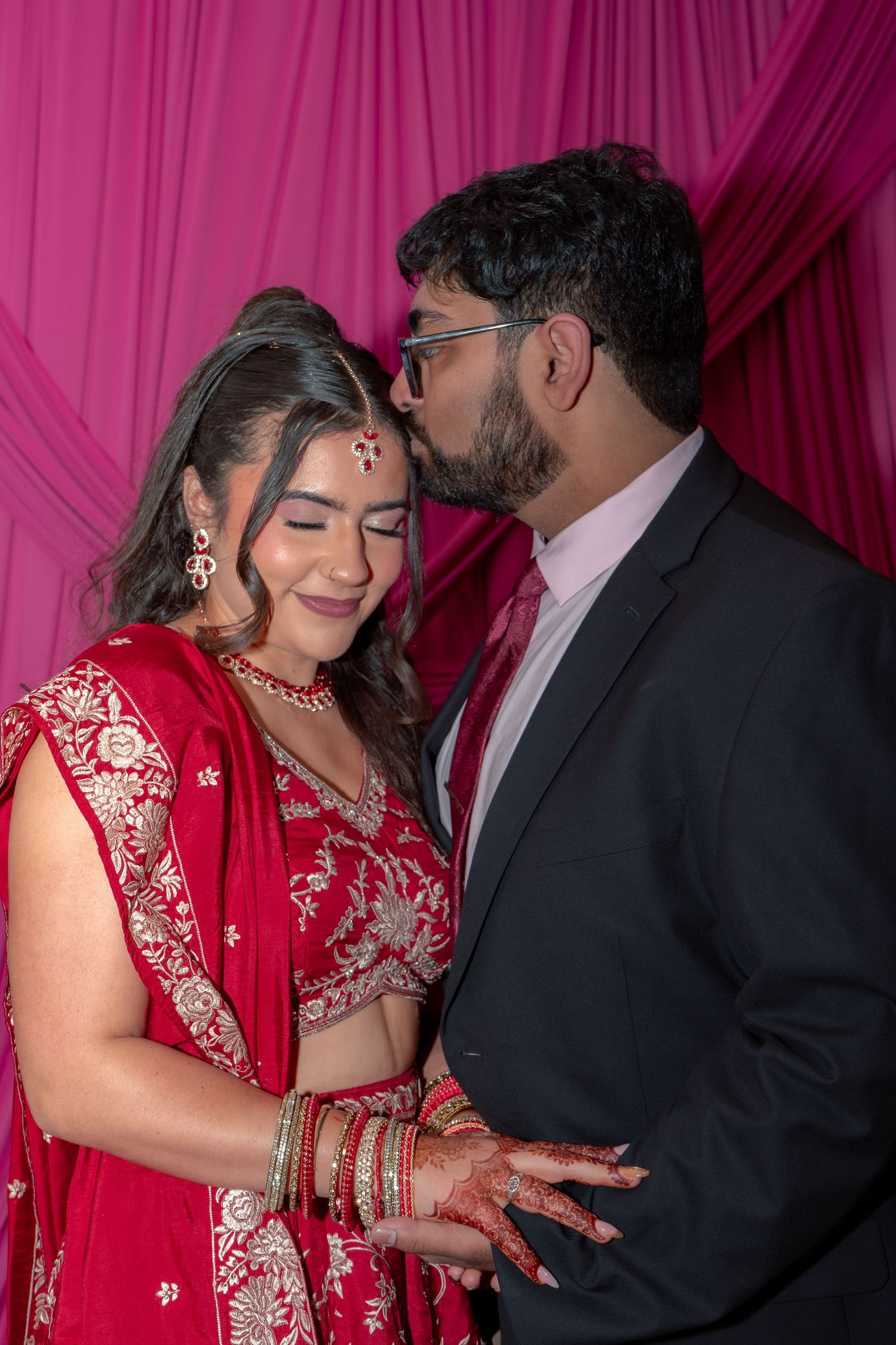 Indian groom in dark suit kissing bride's forehead, bride smiling in red embroidered lehenga with gold jewelry.