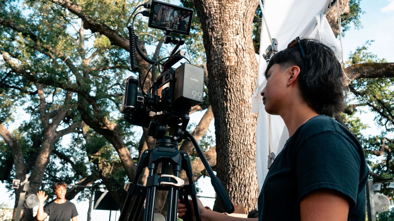 Cinematographer operating a professional film camera on a tripod outdoors near a large tree.
