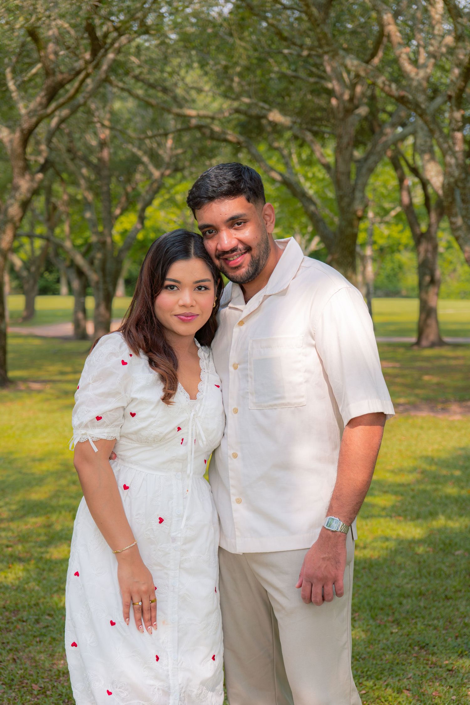 Happy couple in matching white outfits posing together in a lush green park with tree-lined background.