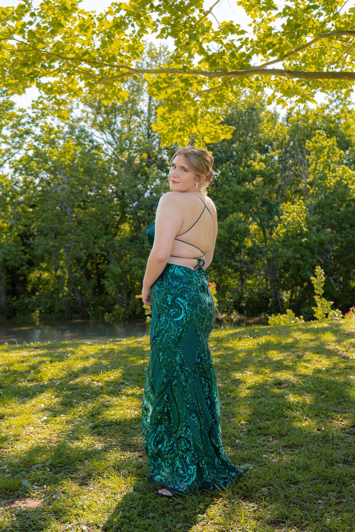 Young woman in a stunning backless emerald green sequin prom gown poses outdoors in a sunlit garden.