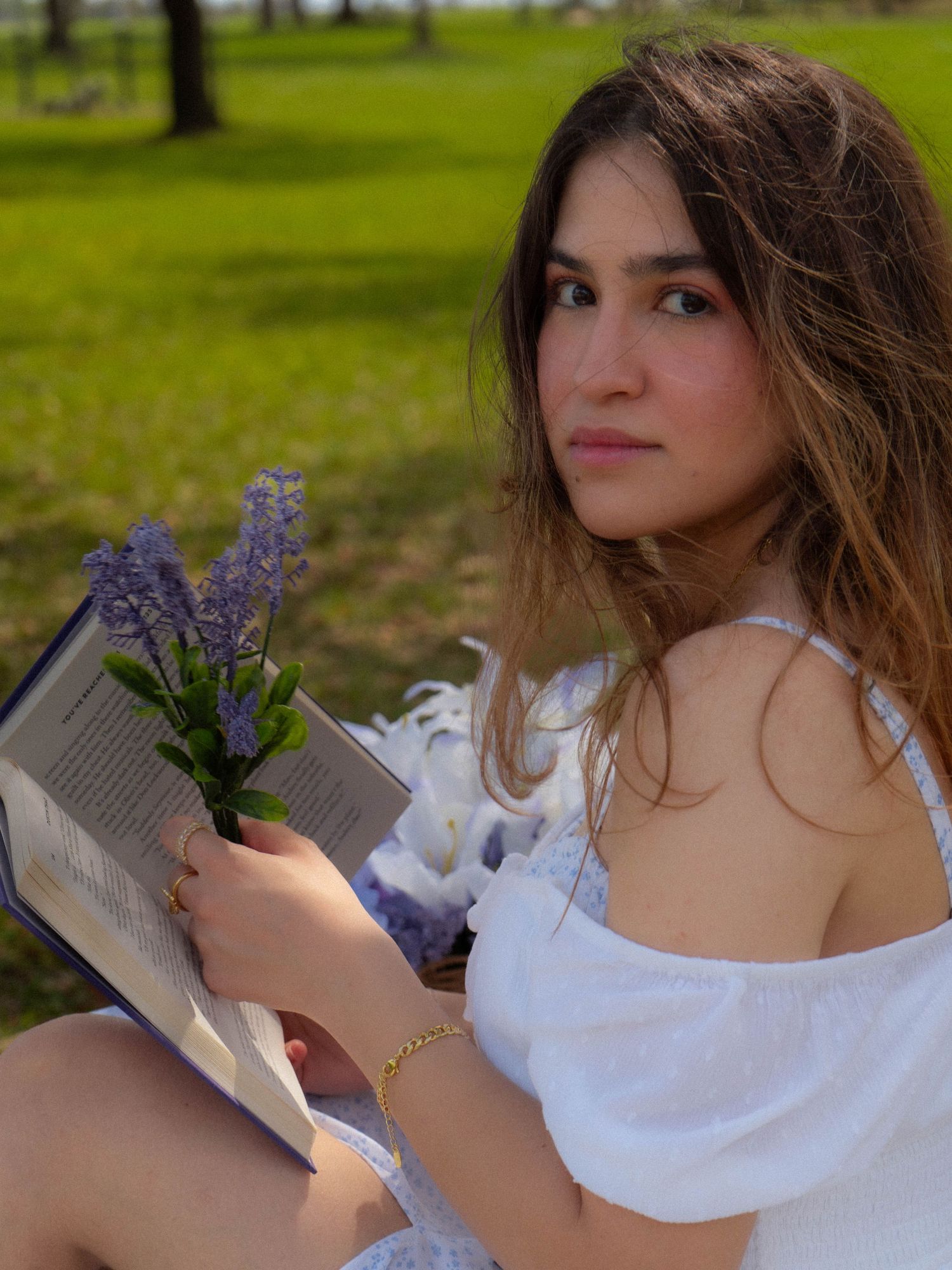 Young woman in white dress holding lavender flowers and open book, seated outdoors in sunny park.
