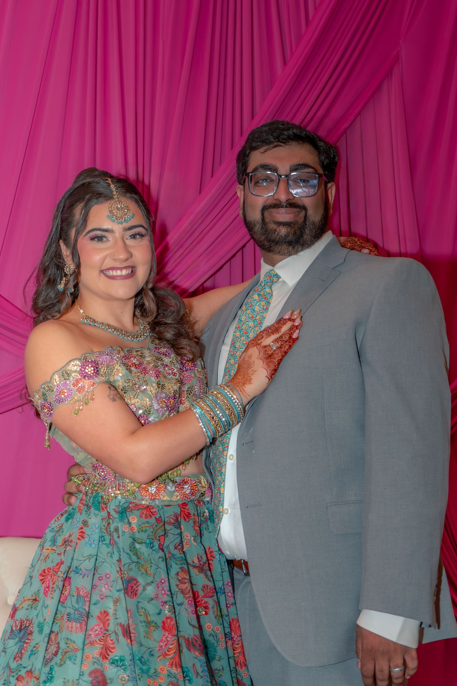 A couple in South Asian wedding attire poses together against a pink draped backdrop at a celebration.