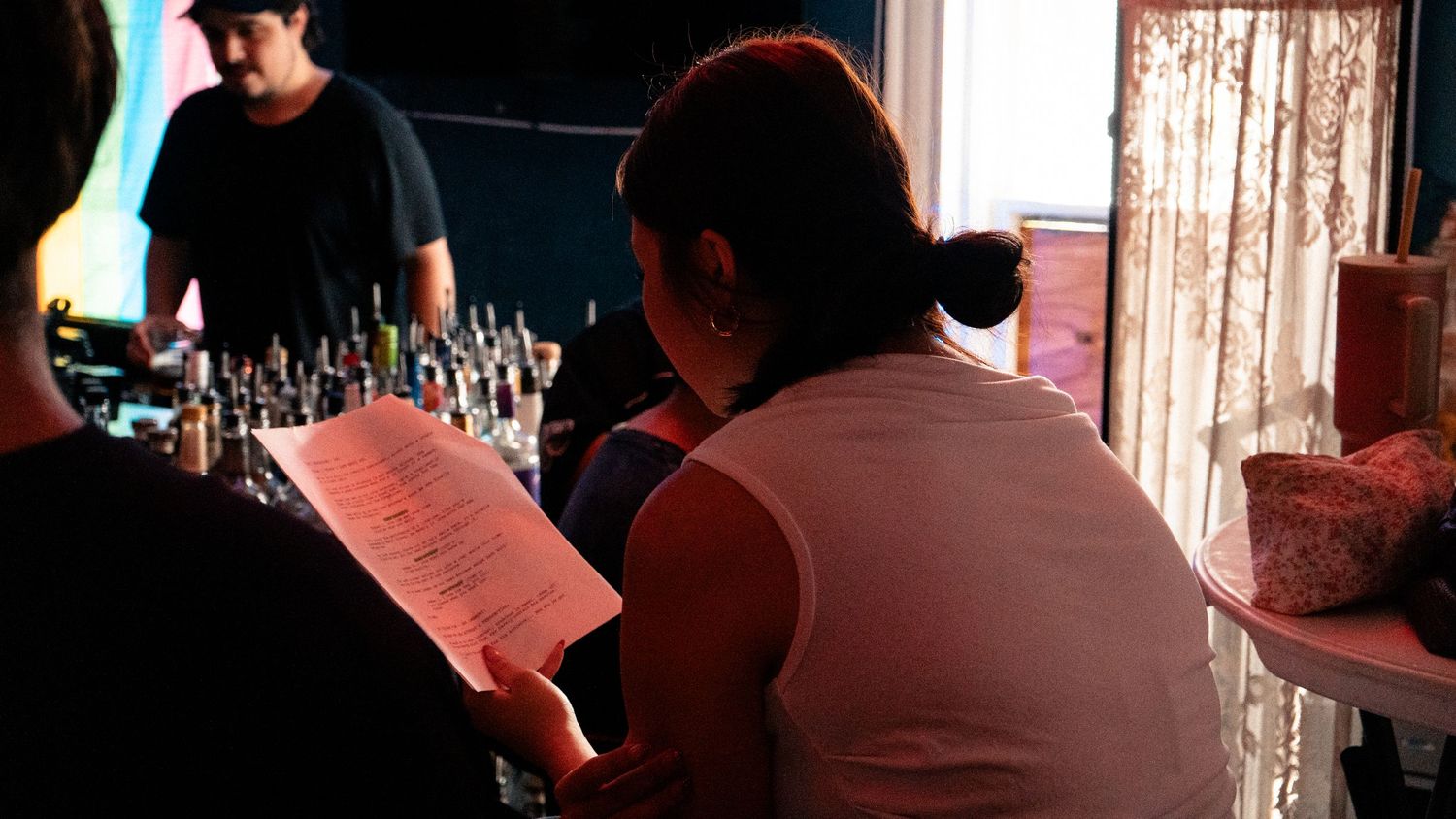 Two people at a dimly lit bar, one reviewing a menu while a bartender works behind a bottle-lined counter.