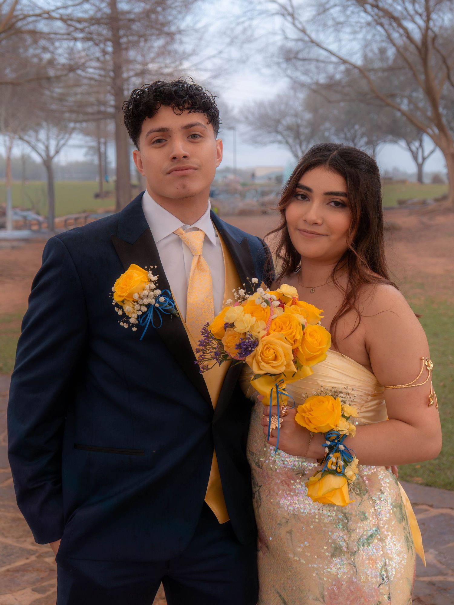 A young couple in prom attire, man in navy suit with yellow tie, woman in gold gown, holding yellow rose bouquet outdoors.