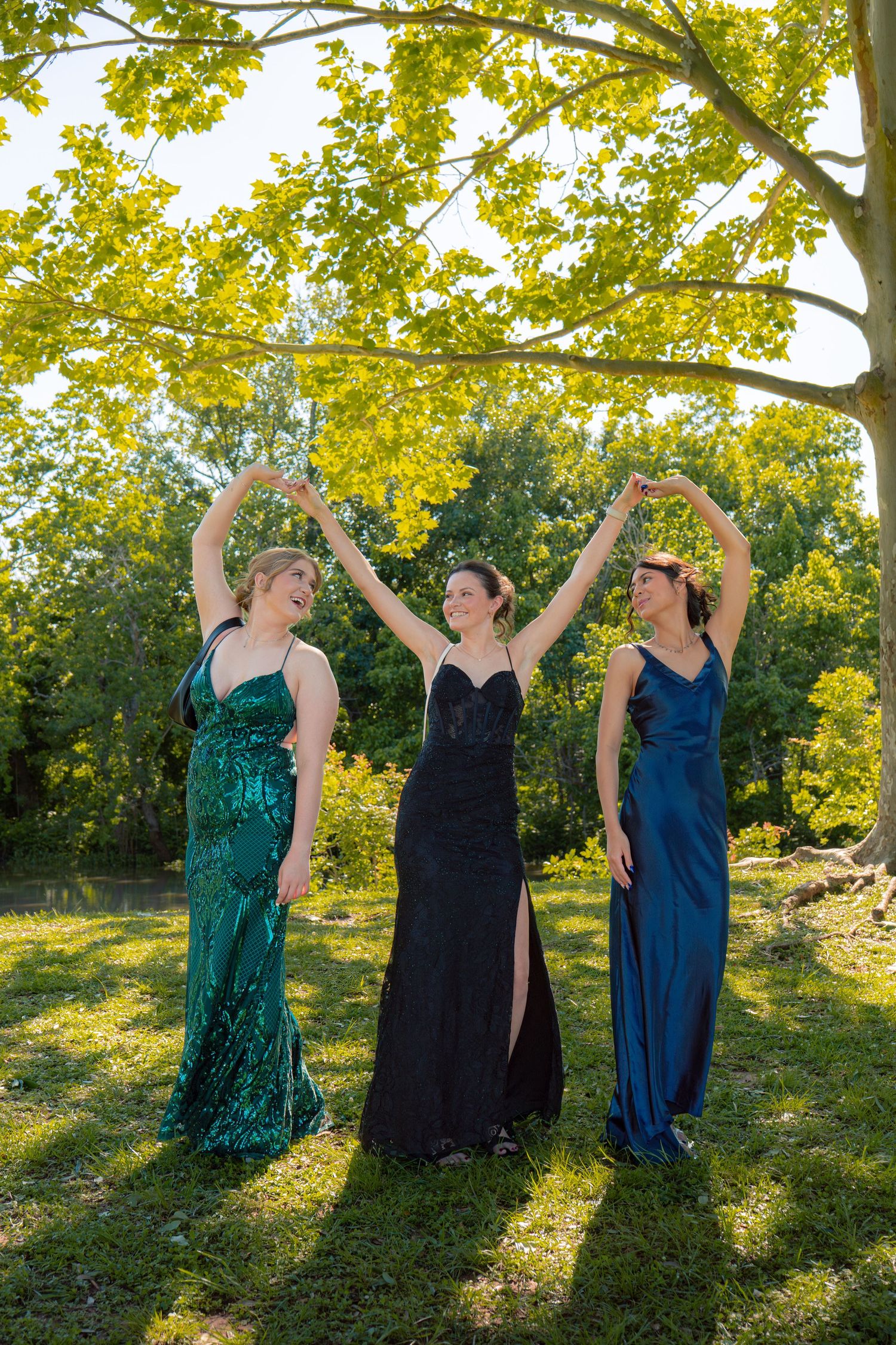 Three young women in elegant prom gowns raise their arms in a park.