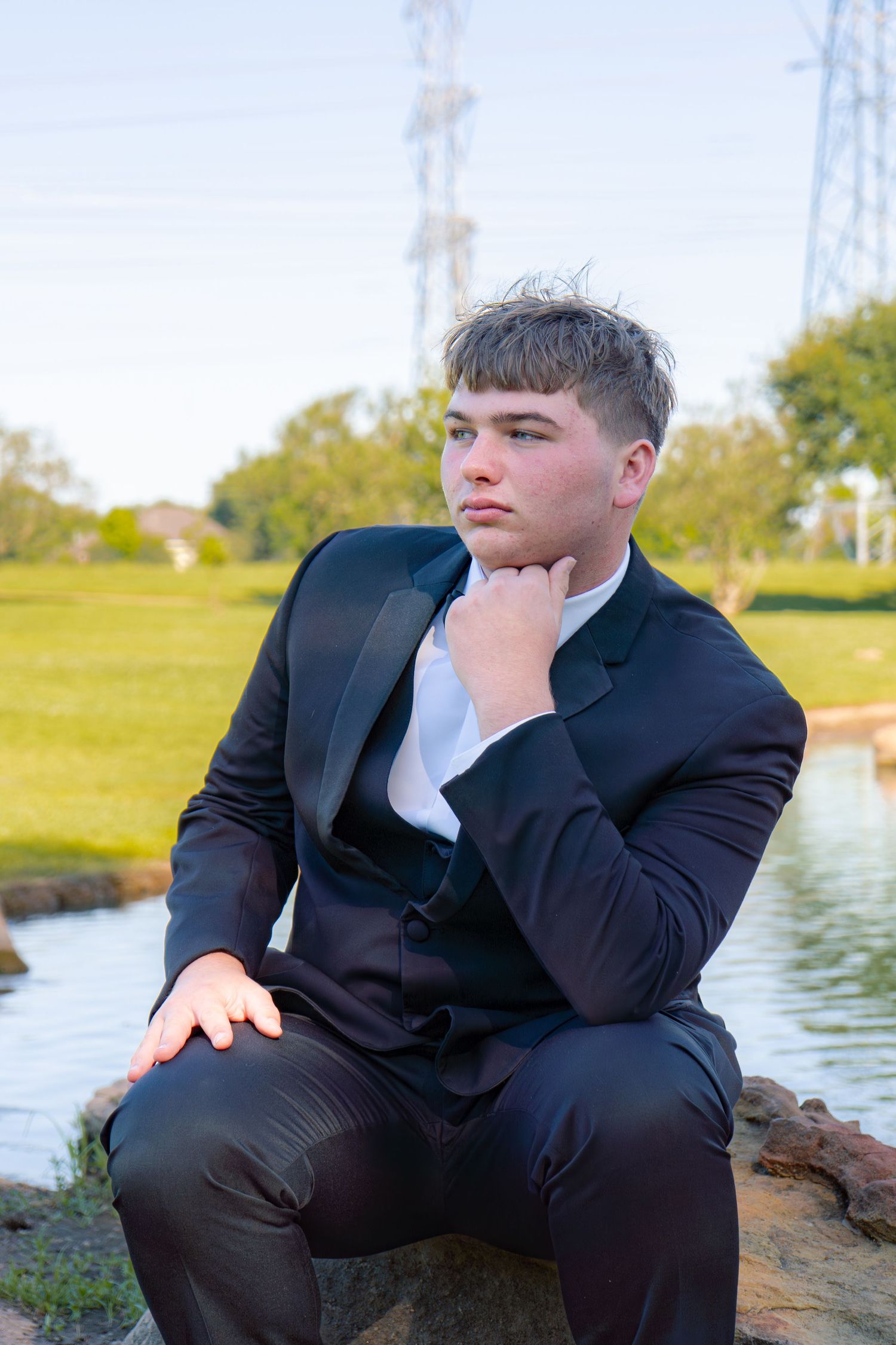 Teenage boy in black tuxedo posing thoughtfully on rocks beside a pond in outdoor park setting.