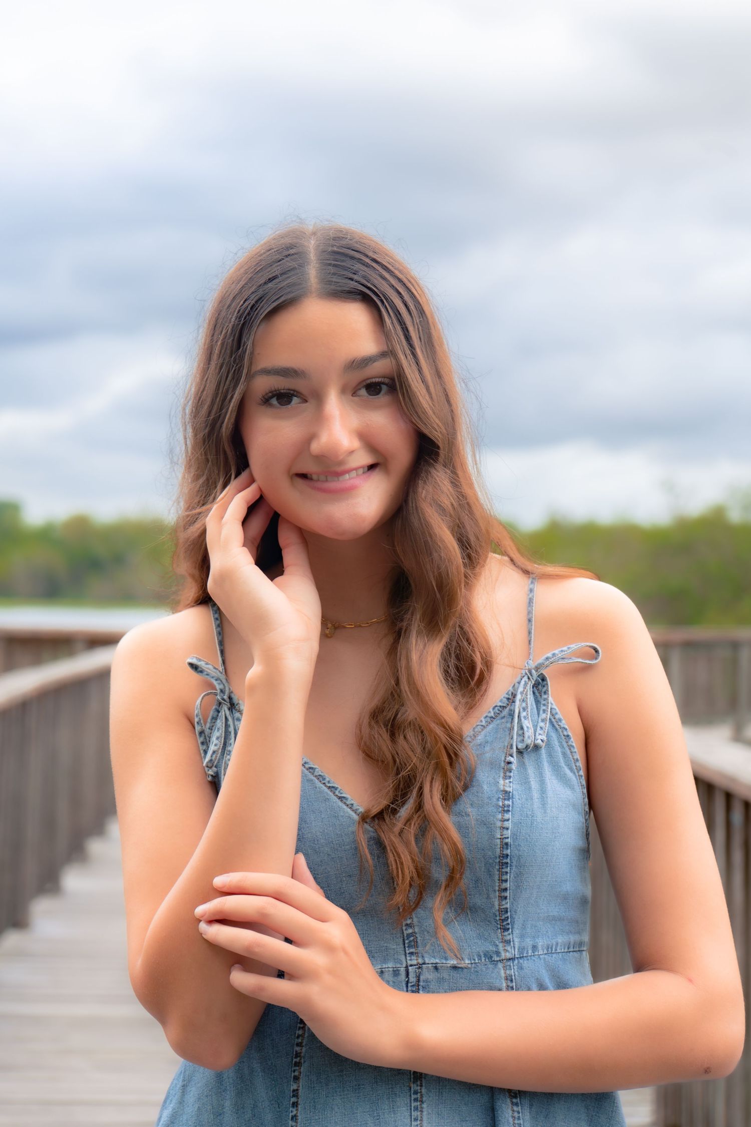 Young woman in denim dress smiling on wooden bridge with cloudy sky backdrop.