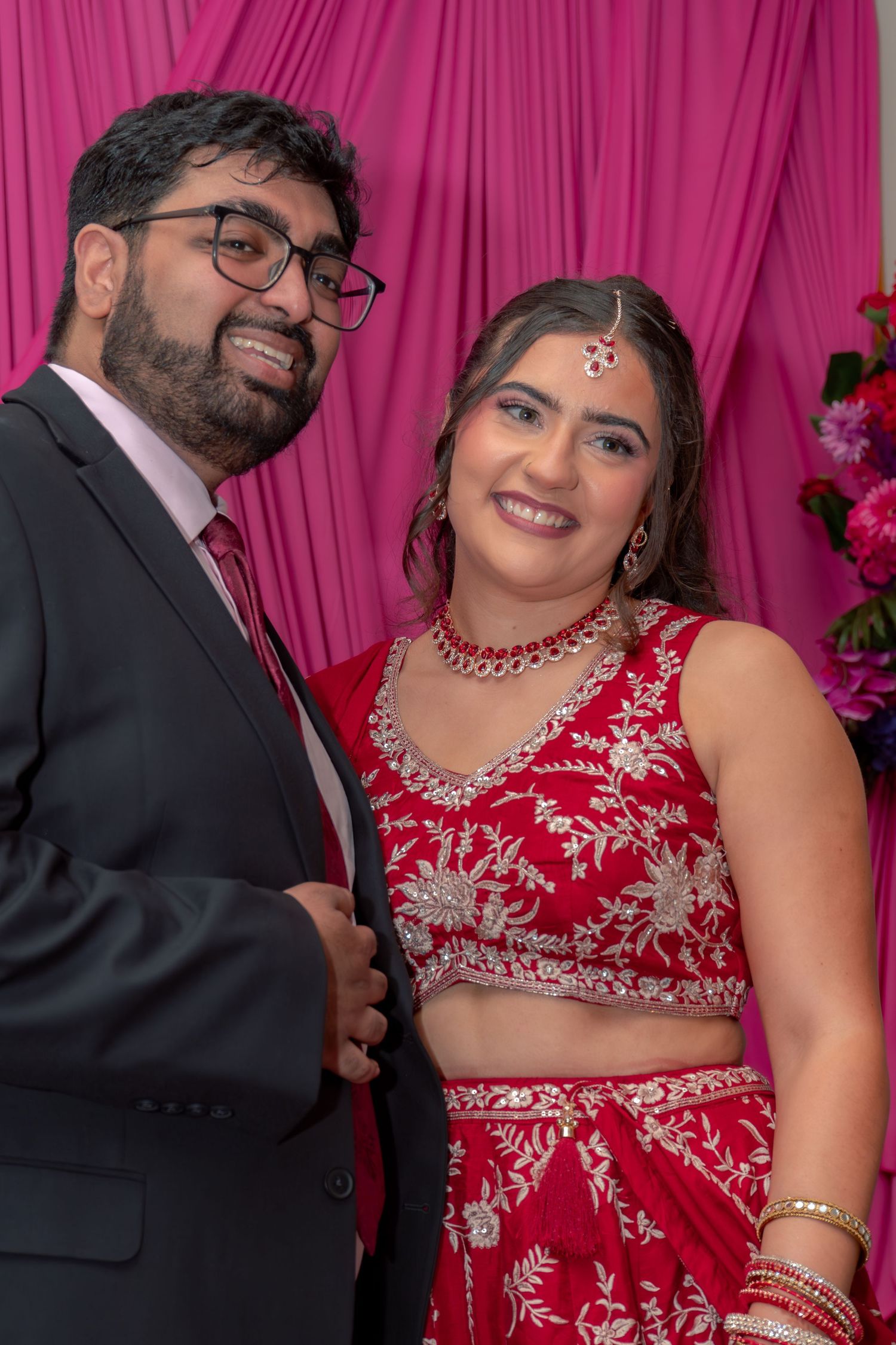 Indian couple in traditional wedding attire, bride in red embroidered lehenga with jewelry, groom in dark suit, pink backdrop.