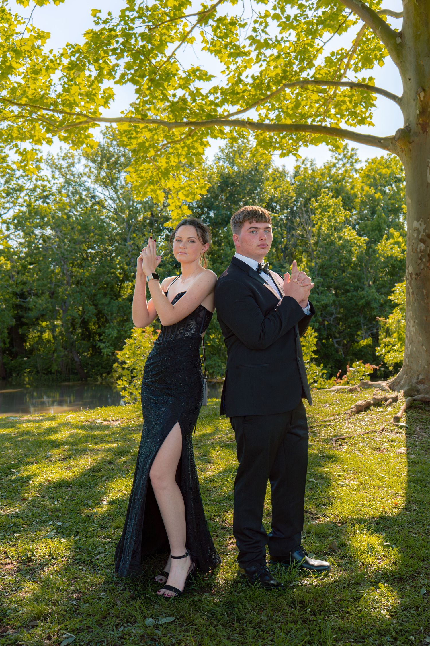 Elegant couple in black formal wear pose back-to-back under a tree at an outdoor prom photoshoot.