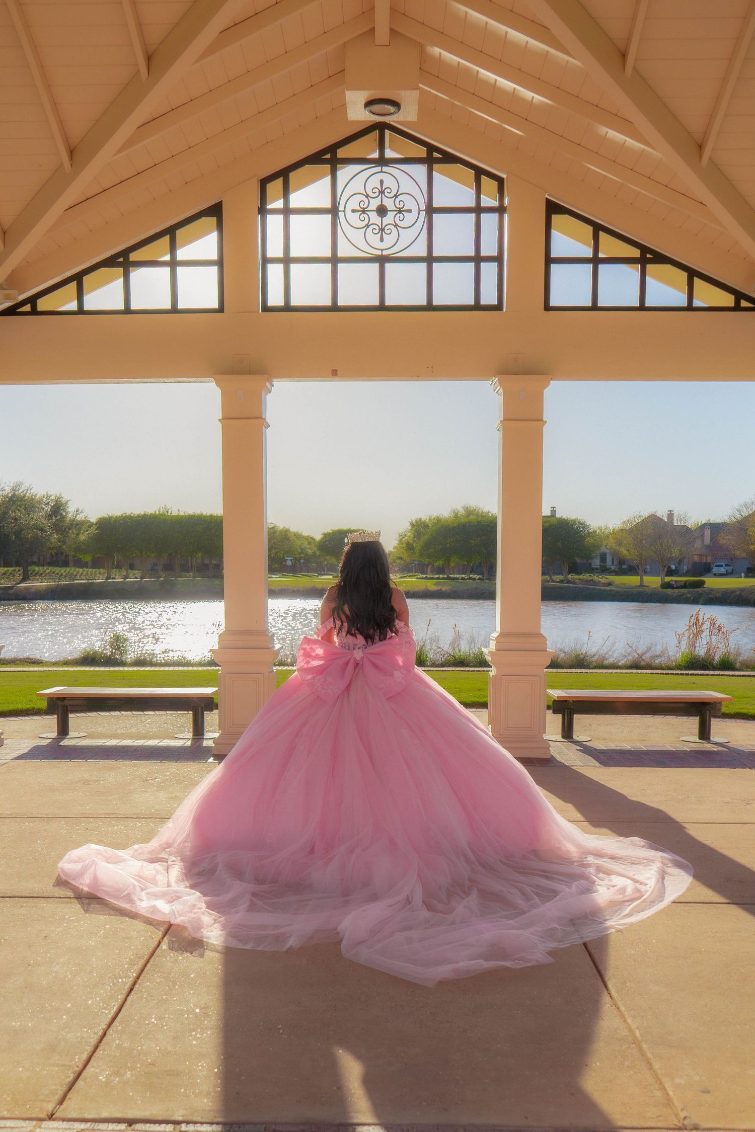 Girl in pink quinceañera ball gown stands in elegant gazebo overlooking serene lake on sunny day.