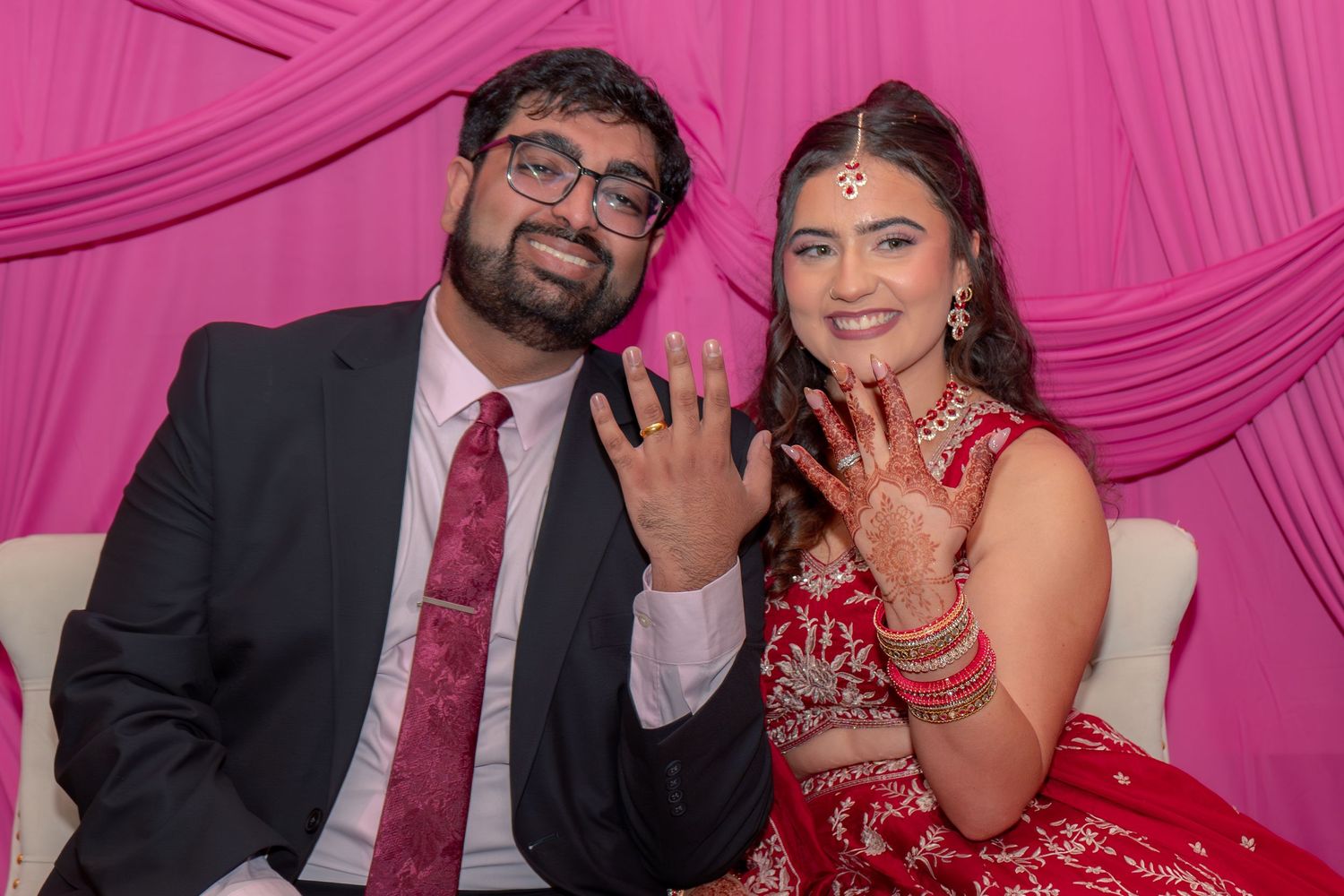 Couple in Indian wedding attire proudly displaying rings with henna hands at engagement ceremony.