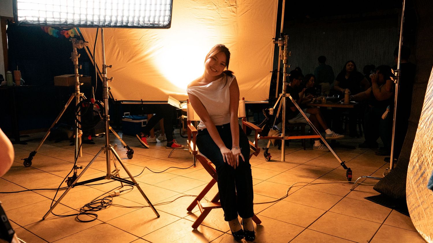 A woman poses confidently on a director's chair on a professional film set with studio lighting equipment.